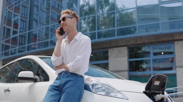 Homem encaracolado moderno elegante em jeans e uma camisa branca no quintal de um complexo residencial falando em um telefone celular, apoiando-se em um carro elétrico enquanto ele está carregando — Vídeo de Stock