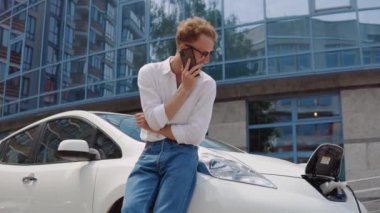 Modern kıvırcık adam elektrikli arabasını bir sitenin bahçesinde şarj ediyor ve cep telefonuyla konuşuyor.