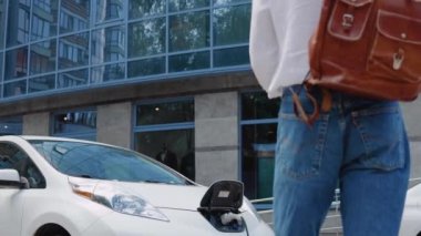 Şık, genç, kıvırcık bir adam omuzunda şık bir şehir çantasıyla elektrikli arabasına doğru yaklaşıyor.