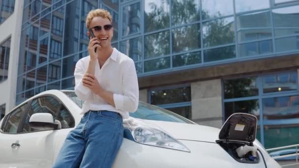 Homem encaracolado moderno elegante carrega seu carro elétrico no quintal de um complexo residencial e fala em um telefone celular — Vídeo de Stock