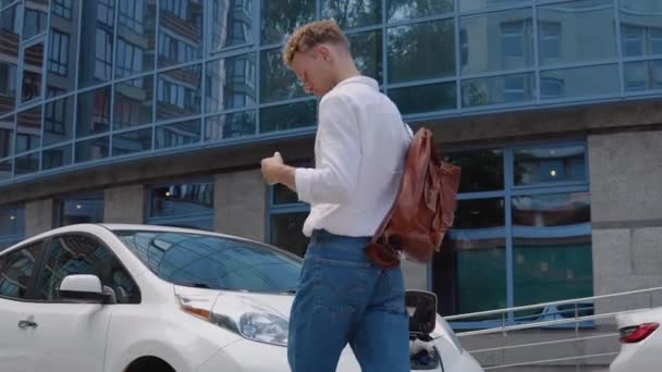 Elegante hombre rizado joven moderno con un elegante maletín de la ciudad en su hombro se acerca a su coche eléctrico mientras se está cargando — Vídeo de stock