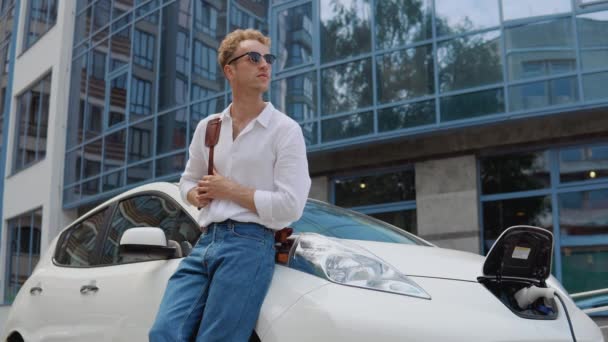 Stijlvolle moderne jonge krullende man met een stijlvolle stad aktetas op zijn schouder zit in de buurt van zijn elektrische auto, terwijl hij laadt — Stockvideo