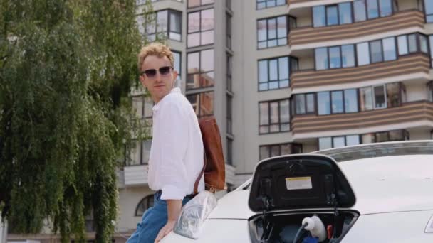 Stylový moderní mladý kudrnatý muž se stylovým městským kufříkem na rameni sedí vedle svého elektrického auta a nabíjí. Detailní pohled na baterii a nabíječku — Stock video