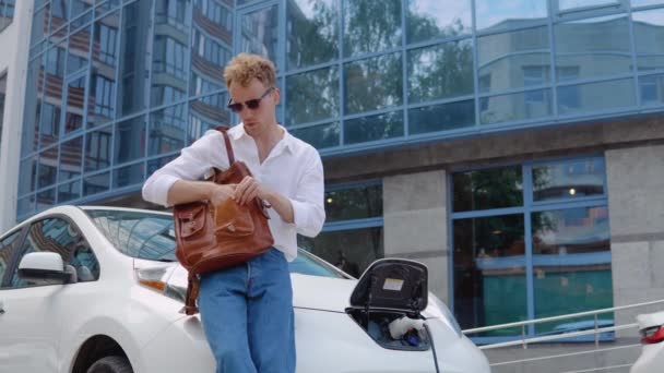 Стильний сучасний молодий кучерявий чоловік зі стильним міським портфелем відходить від свого електромобіля, залишаючи машину на борт — стокове відео