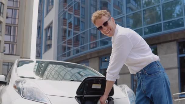 Stylish modern young curly man stands by the battery and holds a connected charger — Stock Video