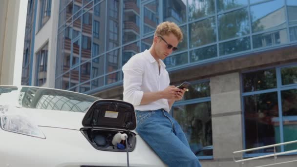 세련 된 현대의 젊고 곱슬곱슬 한 젊은이가 스마트폰으로 돌진하면서 빙글빙글 하는 전기 자동차 근처에서 있다 — 비디오