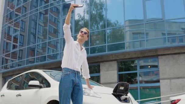 Elegante hombre rizado joven moderno se encuentra cerca de una carga de coche eléctrico y se toma una selfie con su teléfono inteligente — Vídeos de Stock