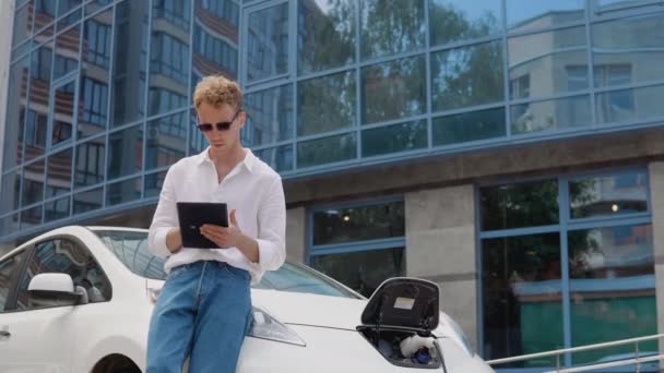 Elegante hombre rizado joven moderno se encuentra cerca de un coche eléctrico de carga y utiliza su tableta — Vídeos de Stock