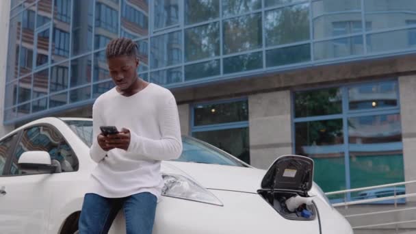 Elegante joven afroamericano de pie cerca de un coche eléctrico de carga y deslizamiento en su teléfono inteligente — Vídeo de stock