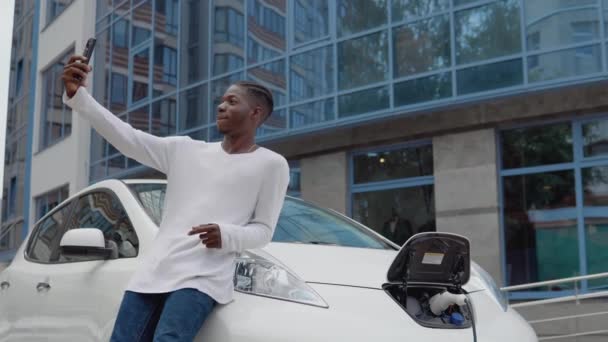 Homem americano africano elegante fica perto de um carregamento de carro elétrico e tira uma selfie com seu smartphone — Vídeo de Stock