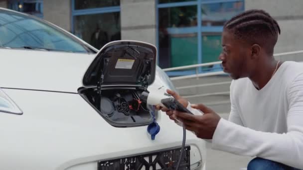 En ung afrikansk amerikansk man ansluter en elektrisk bil till laddaren och justerar processen för att ladda bilbatteriet med hjälp av en mobiltelefon — Stockvideo