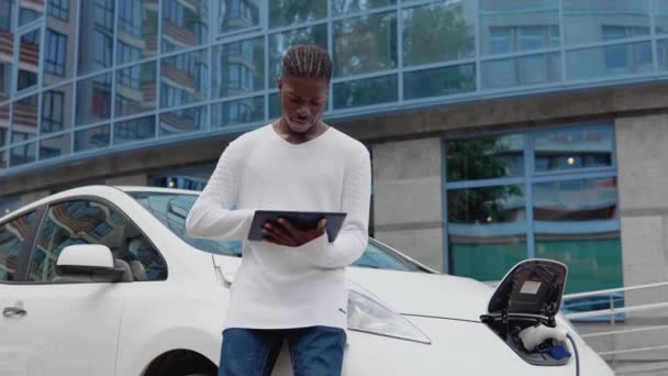 Стильный африканский американец мужчина стоит рядом с электрическим автомобилем заряжая и использует свой планшет — стоковое видео