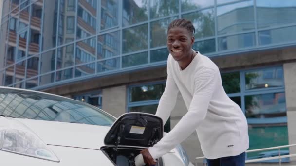 Um jovem afro-americano conecta um carro elétrico ao carregador e olha para a câmera — Vídeo de Stock