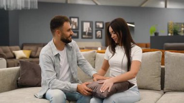 Genç, gülümseyen bir çift mobilya galerisinde rahat bir kanepe ve yastık seçiyor, birlikte oturuyor ve modern dairelerinin alımını tartışıyor ve ortak bir karar veriyorlar.