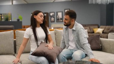 Genç bir adam ve kadın yeni bir kanepe ve yastık seçiyorlar, modern bir mobilya mağazasının vitrininde birlikte otururken alışverişi tartışıyorlar.