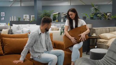 Ev dekorasyonu için alışveriş yapan mutlu çift, büyük bir mobilya galerisinde yeni bir kahverengi kanepe ve uyumlu yastıklar seçiyor. Kadın aksesuarları düzenlerken erkek rahatlığı test ediyor.