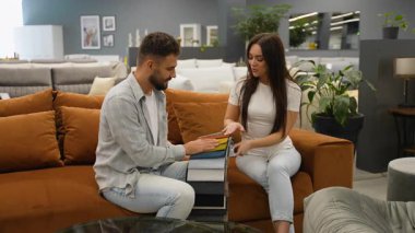 Mutlu genç çift rahat kahverengi bir koltukta otururken örnek bir kitaptan mükemmel döşeme kumaşını seçiyorlar, yeni ev mobilyaları için renkleri ve dokuları dikkatlice seçiyorlar.