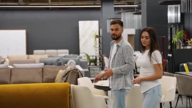 Genç mutlu çift büyük bir mobilya mağazasında yürüyor, modern evleri için yeni bir kanepe ve yemek masası seçiyor ve oturma odası için tasarım tercihlerini tartışıyorlar.