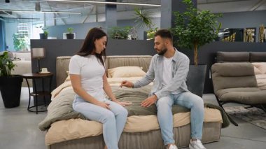 Yeni bir mobilya mağazasında yeni bir yatakta oturan beyaz bir çift, yeni ev seçimlerini tartışırken döşeğe ve yatağa dokunuyor, yatak odası mobilyalarını seçme sürecini sergiliyorlar.