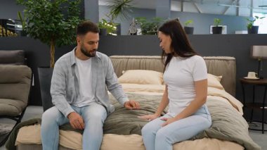 Mobilya mağazasında yeni rahat bir yatakta oturan mutlu genç çift modern evleri için mükemmel yatak odası ve yatağı seçip sohbet ediyorlar.