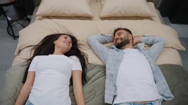 Gülümseyen genç bir çiftin yeni rahat bir yatakta birlikte rahat rahat konuşmalarının, gülmelerinin ve modern, rahat yatak odalarında sakin bir anın tadını çıkarmalarının en tepedeki görüntüsü.