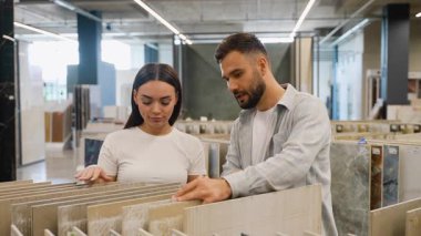 Ev yenileme projeleri için yeni seramik fayansları tartışıp seçen mutlu genç çift, büyük bir modern hırdavat mağazası veya galeride çeşitli örnekleri gözden geçiriyorlar.