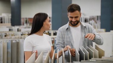 Hırdavatçıdaki seramik zemin ya da duvar fayanslarını seçerken konuşan ve gülümseyen genç beyaz bir çift. Karı koca bir galeride ev yenileme malzemesi alışverişi yapıyorlar.