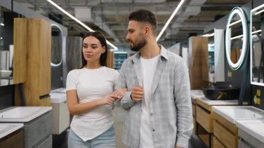 Ev yenileme mağazasında kol kola yürüyen mutlu genç çift, ev yenileme projeleri için yeni banyo lavaboları, mobilyalar ve eşyalar hakkında aktif olarak tartışıyor ve seçiyorlar.