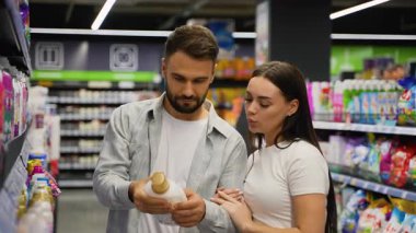 Süpermarkette birlikte alışveriş yapan neşeli genç çift, çamaşır deterjanı ve ev kimyasallarını raftan seçerken sevgi ve mutlulukla konuşuyor ve gülümsüyor.