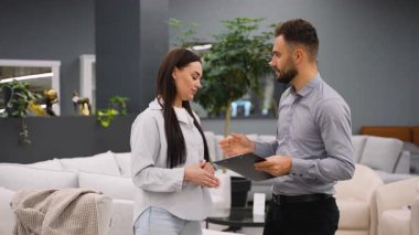 Profesyonel bir erkek mobilya satıcısı genç bir bayan müşteriye tavsiye ediyor, içinde ürün bilgisi olan bir pano gösteriyor. Konsültasyondan sonra tatmin olup, anlaşmaya varmak için el sıkışıyorlar.