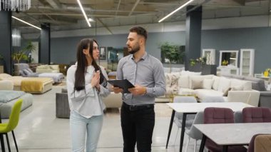 Modern bir mobilya galerisinde kadın müşteriye rehberlik eden bilgili bir satıcı, seçenekleri tartışıyor ve evi için kanepeler, masalar ve sandalyeler arasında yürürken ürünleri işaret ediyor.
