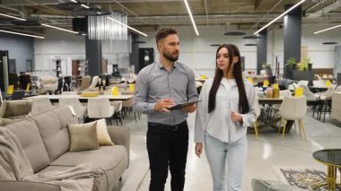 Profesyonel erkek danışman genç bir kadına yeni mobilya seçmesinde yardım ediyor, büyük bir galeride kanepeler ve yataklarla yürüyor, ev geliştirme projesi için tasarım seçeneklerini tartışıyor.