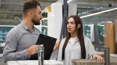 Yeni bir banyo lavabosu ve musluğu seçen genç bir kadın, bir ev geliştirme mağazasında aktif olarak el kol hareketi yaparken, dostane bir satış danışmanı ise profesyonel yardım sağlıyor.