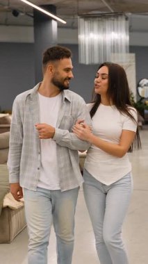 Modern bir mobilya galerisinde el ele yürüyen mutlu genç çift perakende mağazasında birlikte vakit geçirirken evleri için yeni bir kanepe seçip tartışıyorlar.