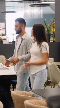 Modern mobilya mağazasında yürüyen mutlu genç çift seçenekleri tartışıyor ve evleri için yeni eşyalar seçiyorlar. Parlak bir galeride alışveriş deneyiminin tadını çıkarıyorlar.