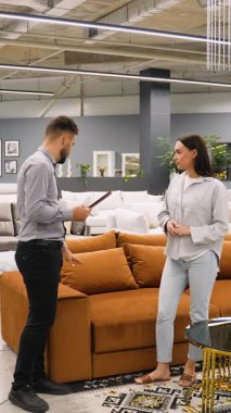 Modern mobilya mağazasında genç bir kadına yeni bir kanepe seçmesinde yardımcı olan profesyonel erkek danışman ürün detaylarını açıklıyor ve ev dekorasyonu seçeneklerini tartışırken notlar alıyor.