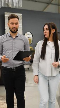 Elinde dosya olan bir erkek danışman genç bir kadına mobilya mağazasında rehberlik ediyor. Satış müdürü ev dekoru seçeneklerini tartışıyor ve müşteriye doğru öğeleri seçmesinde yardımcı oluyor.