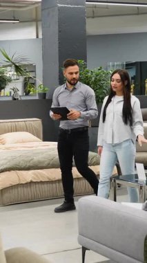 Profesyonel mobilya satıcısı elinde bir pano ve yeni bir kanepe ve yatak seçen genç bir kadına danışıyor ve ona modern ve geniş bir sergi salonundaki çeşitliliği gösteriyor.