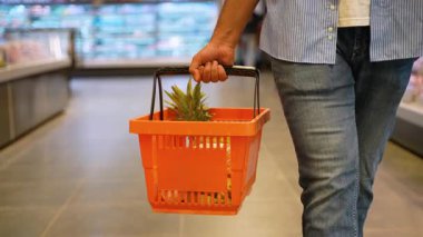 Modern bir marketin koridorunda yürürken turuncu bir alışveriş sepeti ve taze bir ananas taşıyan tanınmamış bir erkek müşteri.