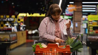 Ciddi genç bir kadın süpermarket koridorunda durup, market alışverişinden sonra uzun alışveriş fişini kontrol ediyor, pahalı toplam fatura ve yükselen fiyatlar için endişeleniyor.