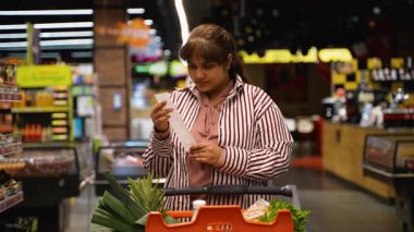 Markette alışveriş makbuzuna bakan stresli genç kadın, yüksek fiyatlar ve gıda enflasyonundan şok olmuş, beklenmeyen toplam maliyetin kendisini hayal kırıklığına uğrattığını hissediyor