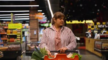 Modern bir süpermarkette alışveriş yapan, içi taze sebzelerle dolu bir arabayı iten ve parlak ışıklı koridorların arasında kameraya güvenle gülümseyen hoş ve iri yarı bir kadın.