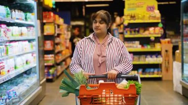 Modern bir süpermarket koridorunda içi taze sebze ve sebze dolu alışveriş arabasını iten düşünceli genç bir kadın sağlıklı bir diyet için raflardan ürünleri dikkatlice seçiyor.