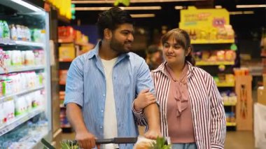 Sevecen genç Hintli çift süpermarket koridorunda alışveriş arabasıyla mutlu bir şekilde yürüyorlar, gülümsüyorlar ve birbirlerine bakıyorlar yemek ve diğer tüketici ürünleri alırken.