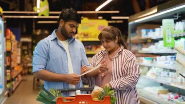 Alışveriş arabalı genç Hintli çift markette ürünlerini seçerken marketin soğutulmuş süt ürünleri reyonunda dikilirken alışveriş listelerini dikkatlice kontrol ediyorlar.