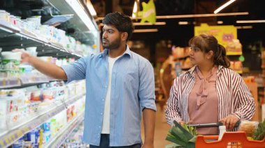 Genç Hintli çift süpermarketin soğutuculu bölümünün yanında durup yoğurdu birlikte seçiyor ve alışveriş arabasına koyuyor ve yiyecekleri hakkında ortak bir karar veriyorlar.