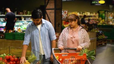 Çekici genç Hintli çift süpermarketin sebze bölümünden taze bir ananas alıp alışveriş arabalarına sağlıklı organik yiyecekler koyuyorlar.