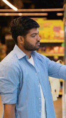 Düşünceli sakallı Hintli adam marketten alışveriş yapıyor, raflardan ürünleri seçiyor ve süpermarkette beklerken bilinçli bir seçim yapmak için etiketleri okuyor.