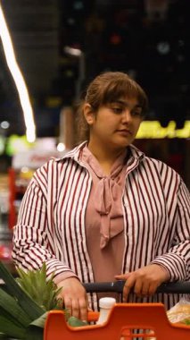 Genç bir kadın, marketin koridoruna dikkatlice bakarken ve alışveriş gezisi sırasında alışverişleriyle ilgili kararlar verirken, içi taze ürünlerle dolu bir alışveriş arabasını itiyor.