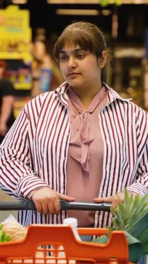 Düşünceli genç bir kadın marketin koridorlarında dolaşırken içi taze ürünlerle dolu bir alışveriş arabasını itiyor. Bir sonraki alışverişini süpermarketin renkli rafları arasında yapmayı düşünüyor.